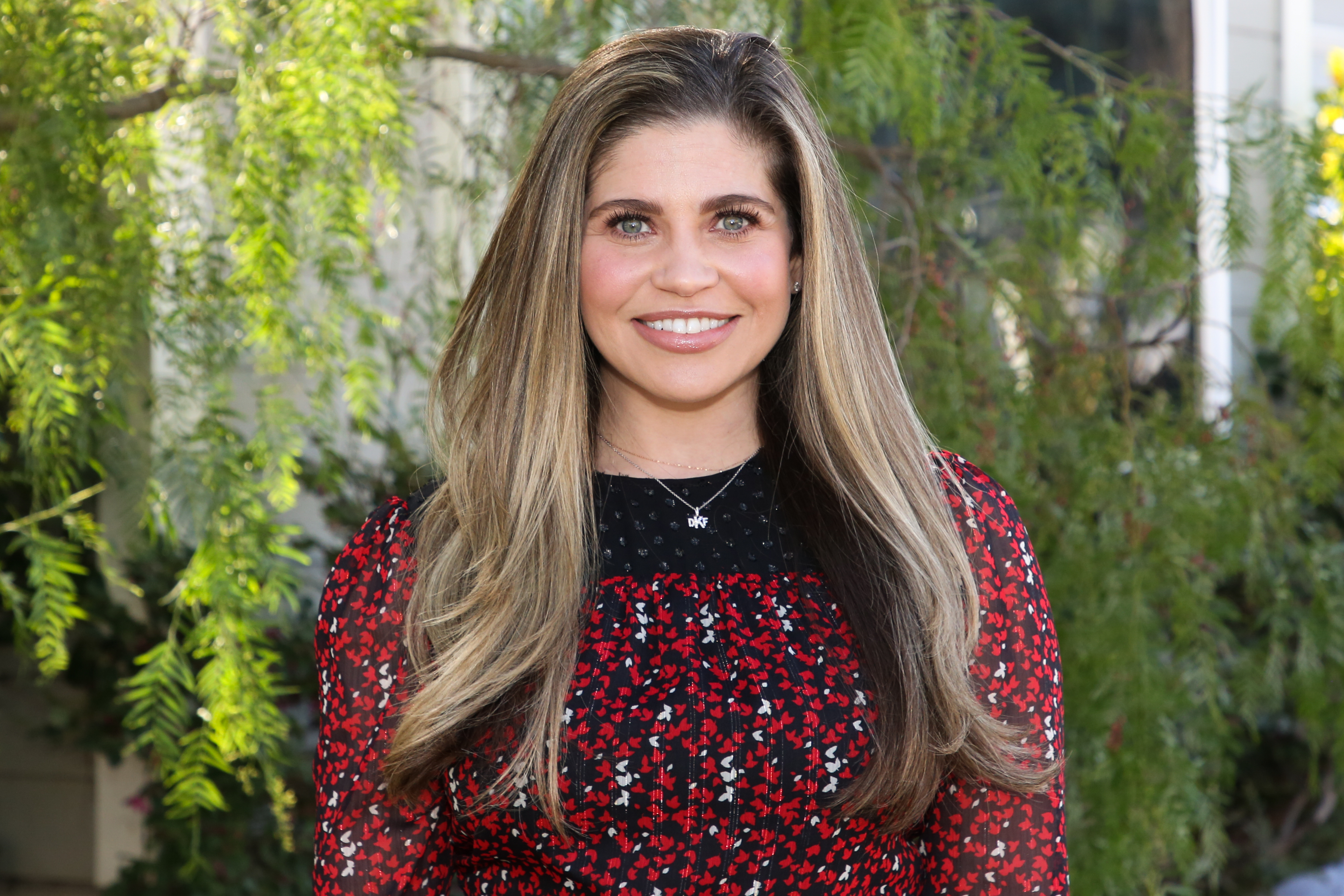 Danielle Fishel smiling, wearing a black dress with a red floral pattern and long sleeves, standing outdoors in front of green foliage