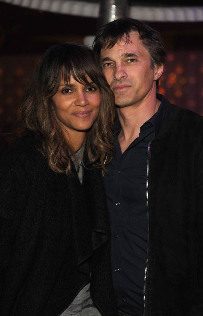 Halle Berry and Olivier Martinez stand closely together, smiling softly at the camera. Halle is wearing a casual outfit, and Olivier is in a dark jacket