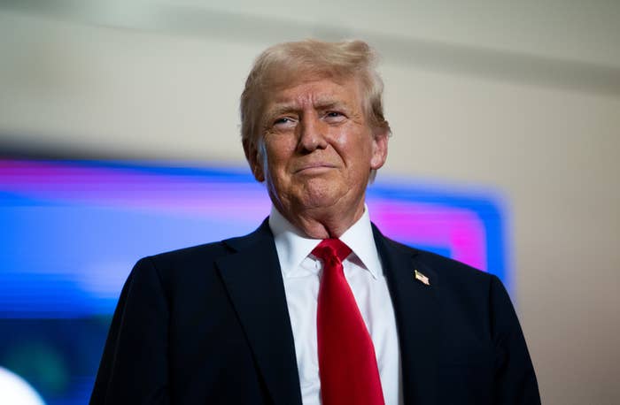 Donald Trump stands in a suit with a white shirt and red tie, looking forward with a slight smile. Background features a blue and purple screen