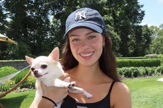 Nicola Peltz Beckham in a black tank top and a New York Yankees cap holds a small white Chihuahua in an outdoor garden.