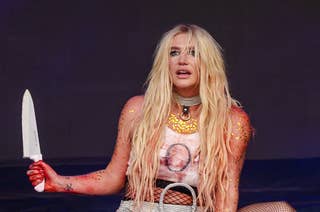 Kesha performs on stage, crouching with a prop knife. She wears a graphic tank top, metallic skirt, fishnet stockings, and Converse shoes