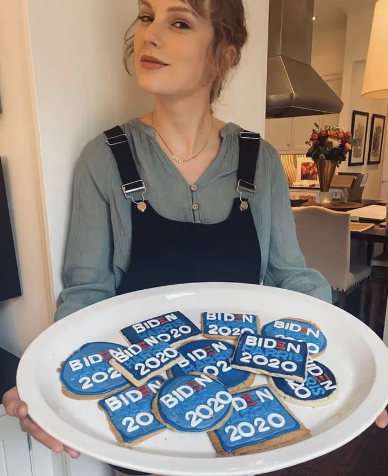 Taylor Swift holding a platter of cookies with &quot;Biden 2020&quot; icing in a kitchen setting. She is wearing a light shirt under a dark pinafore